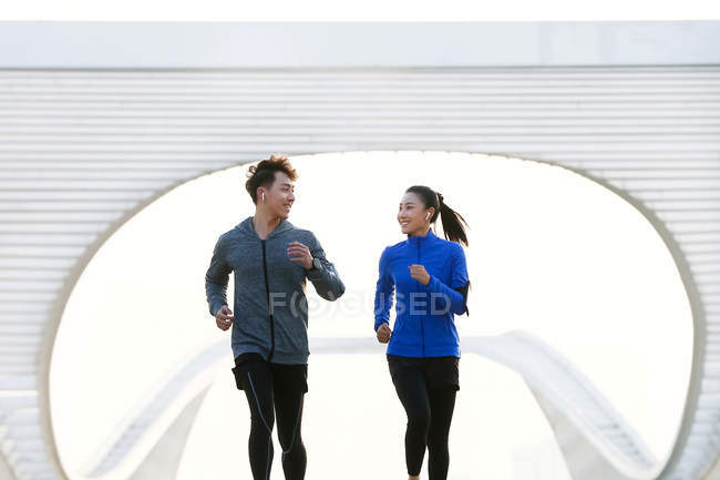 Jóvenes atletas masculinos y femeninos sonriendo entre sí y corriendo juntos en el puente - foto de stock