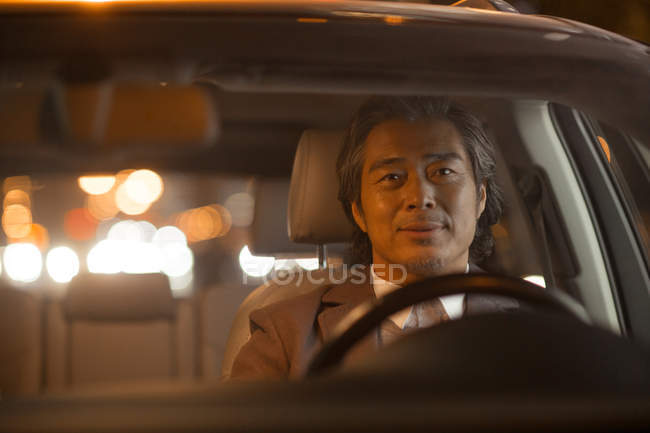 Зрілий азіатський чоловік водить машину і посміхається на камеру вночі — стокове фото