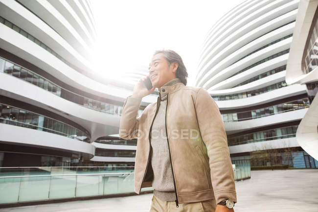 Vue à angle bas d'un homme d'affaires mature souriant parlant par smartphone près d'un immeuble de bureaux moderne — Photo de stock