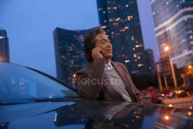 Усміхнений азіатський чоловік спирається на машину і розмовляє по смартфону в нічному місті — стокове фото