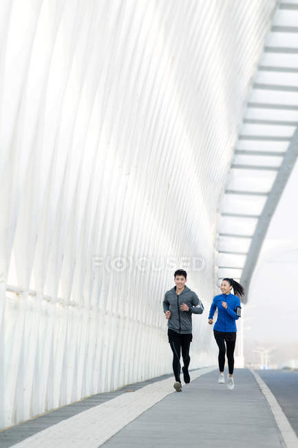 Young asian man and woman in sportswear running together on modern bridge — Stock Photo