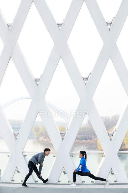 Вид сбоку спортивной молодой пары, растягивающейся во время тренировки на мосту — стоковое фото