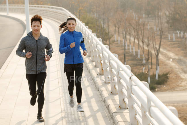 High angle view of sporty young asian couple running on bridge and smiling at camera — Stock Photo