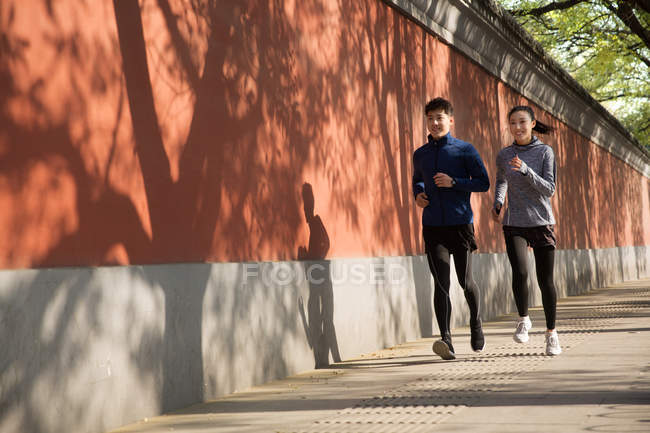 Полный вид молодой азиатской пары в спортивной одежде, улыбающейся и бегающей вместе по улице — стоковое фото