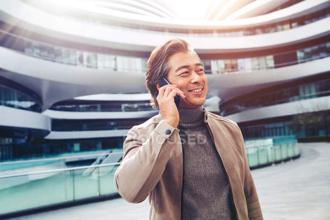 Sonriente asiático hombre de negocios hablando por teléfono inteligente y mirando moderno edificio de oficinas - foto de stock