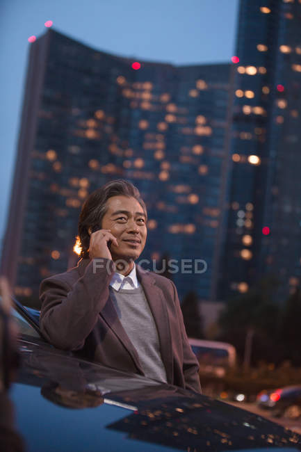 Smiling mature businessman leaning at car and talking by smartphone in night city — Stock Photo