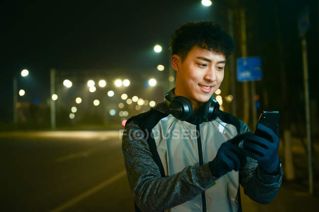 Sonriente asiático hombre en auriculares y ropa deportiva usando smartphone en la noche de la ciudad - foto de stock
