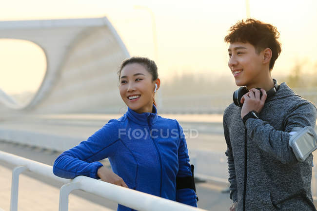 Усміхнена молода азіатська пара в спортивному одязі, стоячи на мосту і дивлячись далеко після тренування — стокове фото