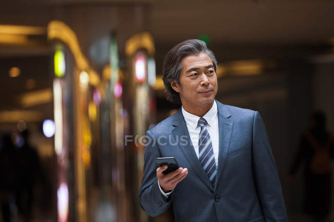 Vorderseite von selbstbewussten reifen asiatischen Geschäftsmann in formeller Kleidung hält Smartphone und schaut weg — Stockfoto