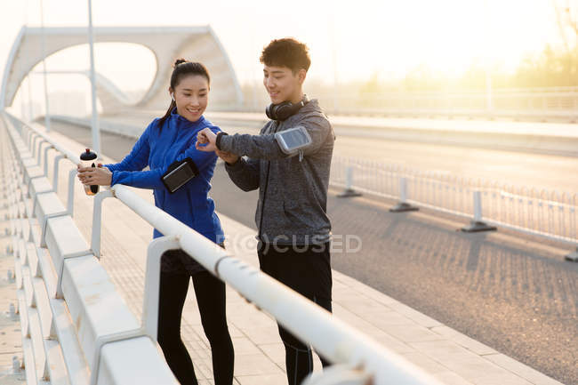 Улыбающаяся молодая пара проверяет наручные часы, стоя вместе на мосту после тренировки — стоковое фото