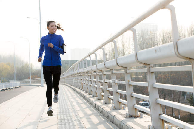 Vue pleine longueur de la jeune femme souriante en vêtements de sport courir sur le pont et regarder loin — Photo de stock