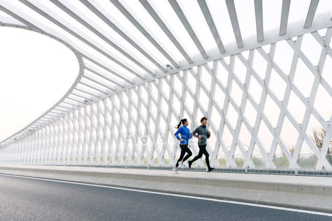 Vista lateral de jóvenes atletas masculinos y femeninos asiáticos corriendo juntos en puente - foto de stock
