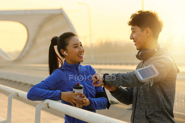 Atlético joven asiático pareja sonriendo uno al otro y chequeando smartwatch en puente en la mañana - foto de stock