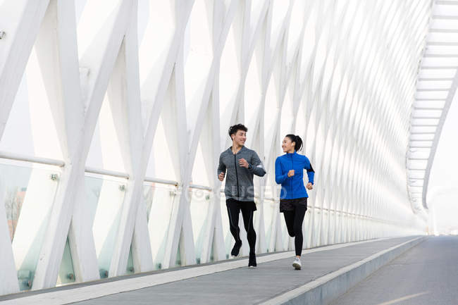 Sportliches junges Paar lächelt einander an und rennt gemeinsam auf Brücke — Stockfoto