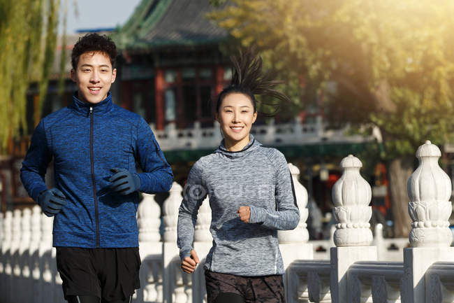 Feliz joven asiático pareja sonriendo en cámara y trotando juntos en la calle - foto de stock