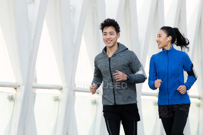 Smiling sporty young couple in sportswear running together on bridge — Stock Photo