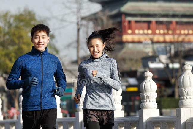 Vista frontal de jóvenes atletas asiáticos sonriendo a la cámara y trotando juntos en la calle - foto de stock