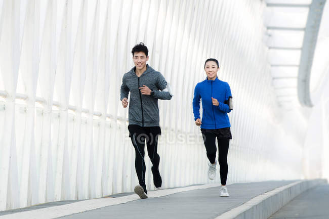 Full length view of smiling young asian man and woman in sportswear running together on bridge — Stock Photo