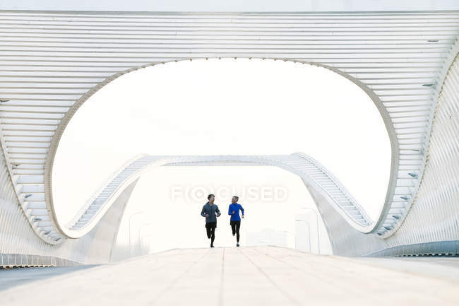 Ganzkörperansicht eines jungen asiatischen Paares in Sportbekleidung, das sich gegenseitig anlächelt und gemeinsam auf einer Brücke läuft — Stockfoto