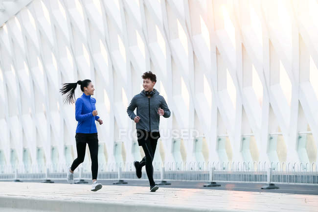 Sporty young asian couple smiling each other and running together on bridge — Stock Photo