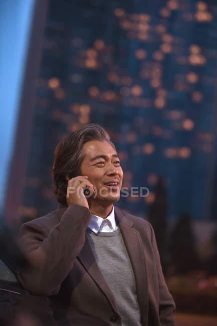 Щасливий зрілий азіатський чоловік розмовляє по смартфону і дивиться в нічне місто — стокове фото
