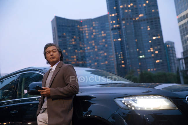 Faible angle de vue de mature asiatique homme debout avec smartphone à côté de la voiture et regardant loin — Photo de stock