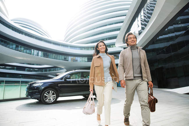 Alegre asiático empresarios caminando cerca de aparcamiento en moderno centro de negocios - foto de stock