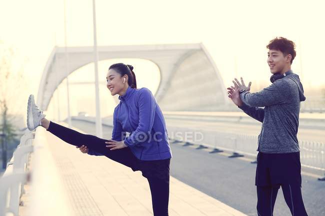 Smiling young asian couple in sportswear stretching during workout on bridge — Stock Photo