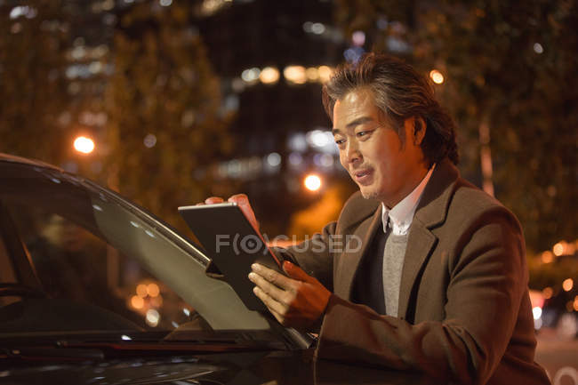 Улыбающийся зрелый азиат, стоящий рядом с автомобилем и использующий цифровой планшет ночью — стоковое фото