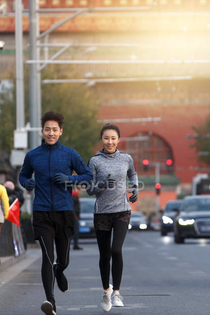Feliz deportivo joven asiático pareja sonriendo a cámara y corriendo juntos en la calle - foto de stock