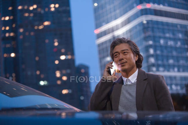 Smiling mature asian man standing beside car and talking by smartphone at night — Stock Photo