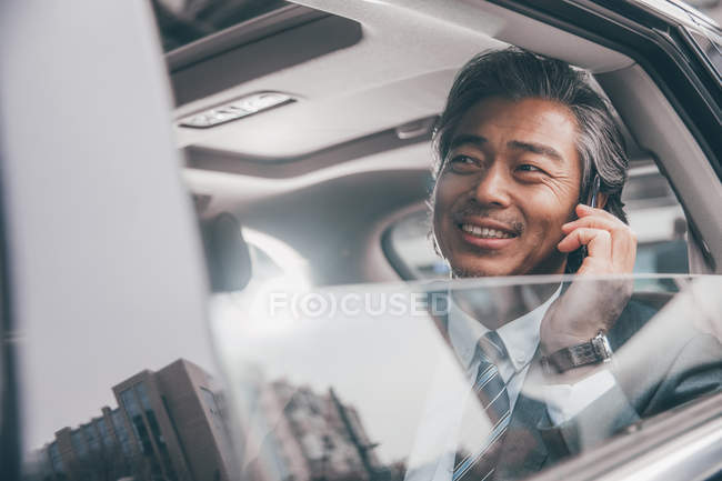 Sorridente uomo d'affari asiatico seduto in auto e parlando con smartphone, messa a fuoco selettiva — Foto stock