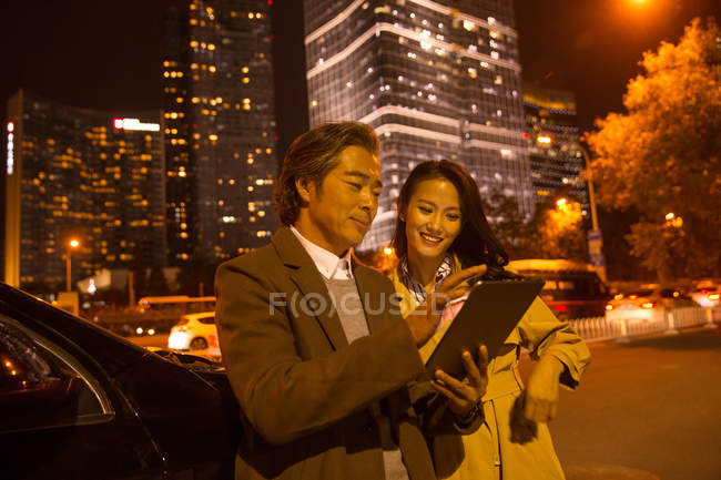 Feliz pareja asiática mirando tableta cerca de coche por la noche - foto de stock