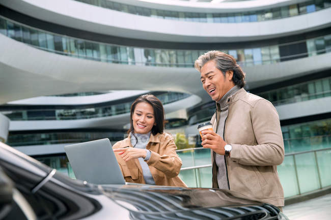 Cheerful businesspeople using laptop on car hood near modern business center — Stock Photo