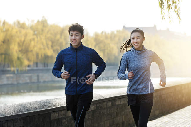 Sonriendo joven asiático hombre y mujer jogging juntos cerca de river - foto de stock
