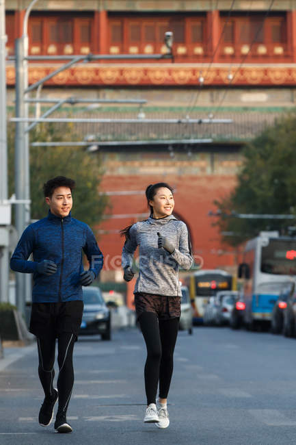 Vista frontal de sonriente joven atlético asiático pareja corriendo juntos y mirando hacia la calle - foto de stock