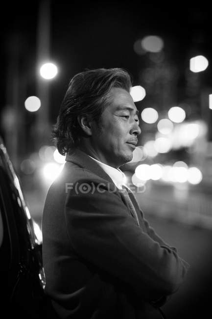 Black and white image of serious mature asian businessman standing near in night city, side view — Stock Photo