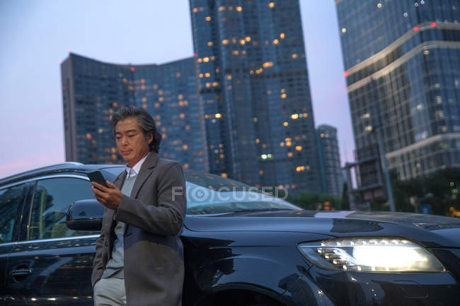 Mature asian man standing beside car and using smartphone — Stock Photo