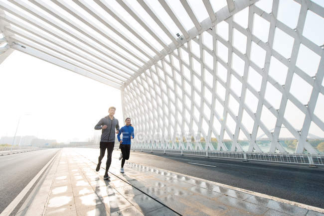 Full length view of young asian man and woman in sportswear running together on bridge — Stock Photo