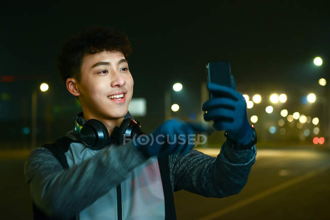 Sonriente asiático hombre en auriculares y ropa deportiva tomando selfie con smartphone en la noche de la ciudad - foto de stock