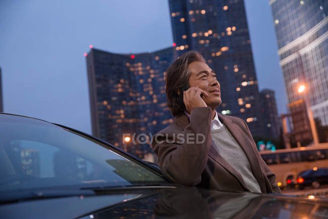 Lächelnd reifer asiatischer Mann lehnt am Auto und spricht mit dem Smartphone in der Nacht Stadt — Stockfoto