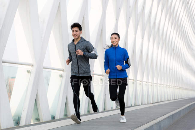 Улыбающаяся спортивная молодая азиатская пара бегущая вместе по мосту — стоковое фото