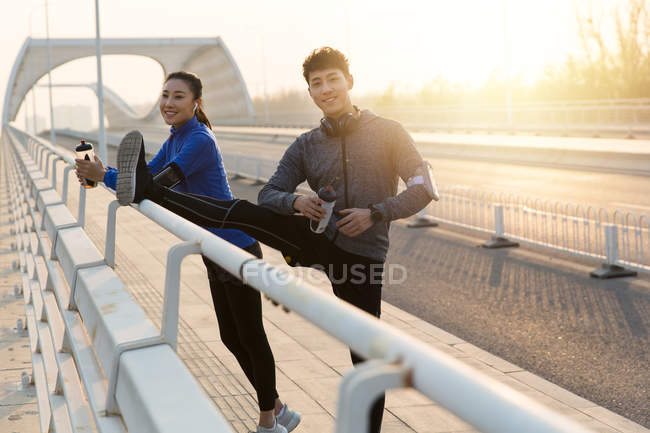 Giovani corridori sorridenti che tengono bottiglie d'acqua e si allungano insieme sul ponte — Foto stock