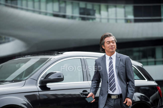 Lächelnd reifer asiatischer Geschäftsmann in offizieller Kleidung, der sein Smartphone neben dem Auto hält — Stockfoto