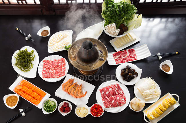 Vista de ángulo alto de platos con varios ingredientes, palillos y olla caliente de cobre, concepto de plato de roce - foto de stock