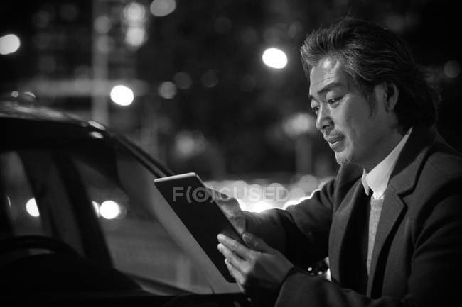 Schwarz-Weiß-Bild fokussierter reifer asiatischer Geschäftsmann, der in der Nähe des Autos steht und nachts ein digitales Tablet benutzt — Stockfoto