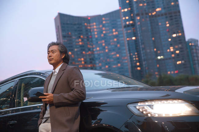 Стомлений азіатський чоловік спирається на машину і тримає смартфон — стокове фото