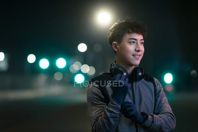 Smiling young asian runner in headphones standing on street and looking away in night city — Stock Photo