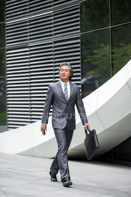 Full length view of smiling asian businessman in formal wear holding briefcase and walking on street — Stock Photo