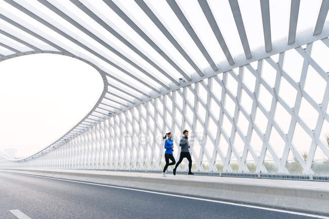 Полный обзор молодой пары бегунов в спортивной форме, тренирующихся вместе на современном мосту — стоковое фото